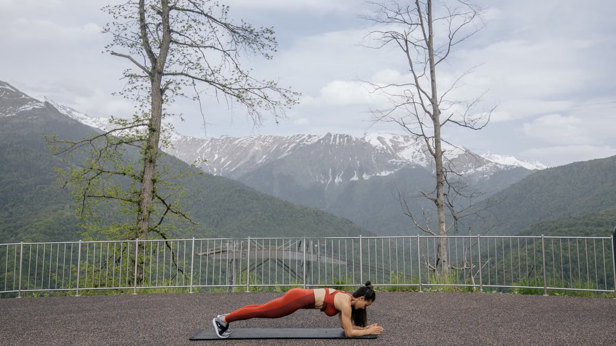 A woman doing a plank exercise on a mat outdoors with a scenic mountain view in the background.